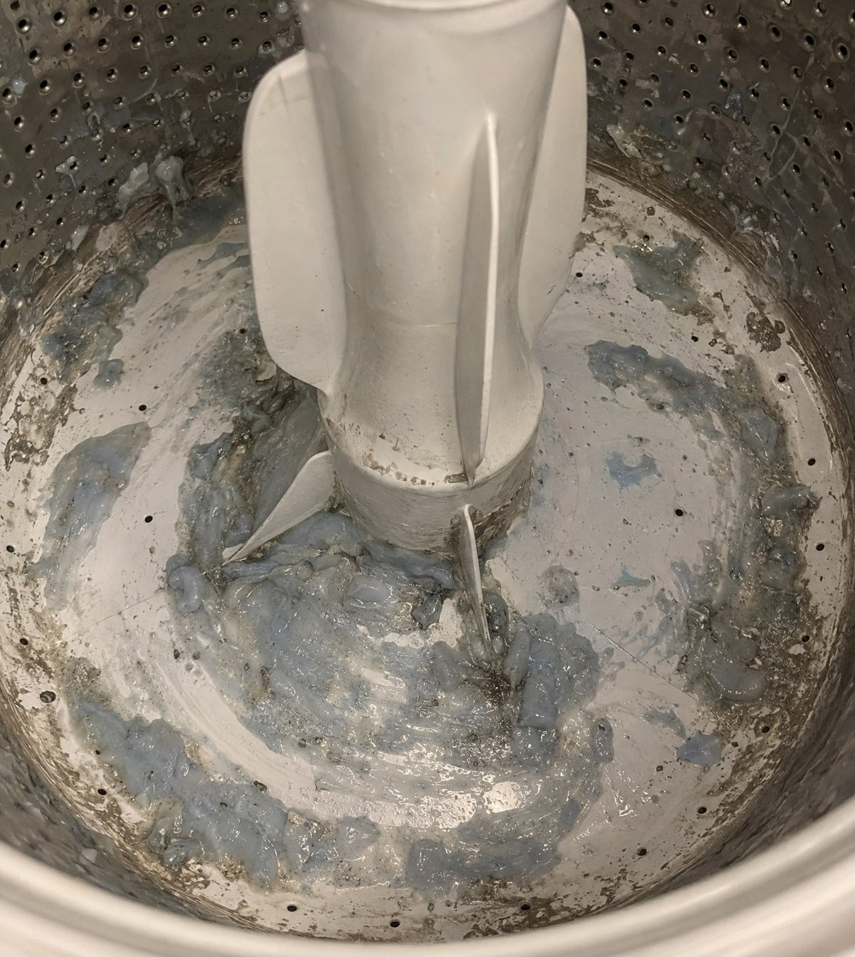 Illustration of a top-loading washer tub floor coated in gray detergent residue around the agitator, before